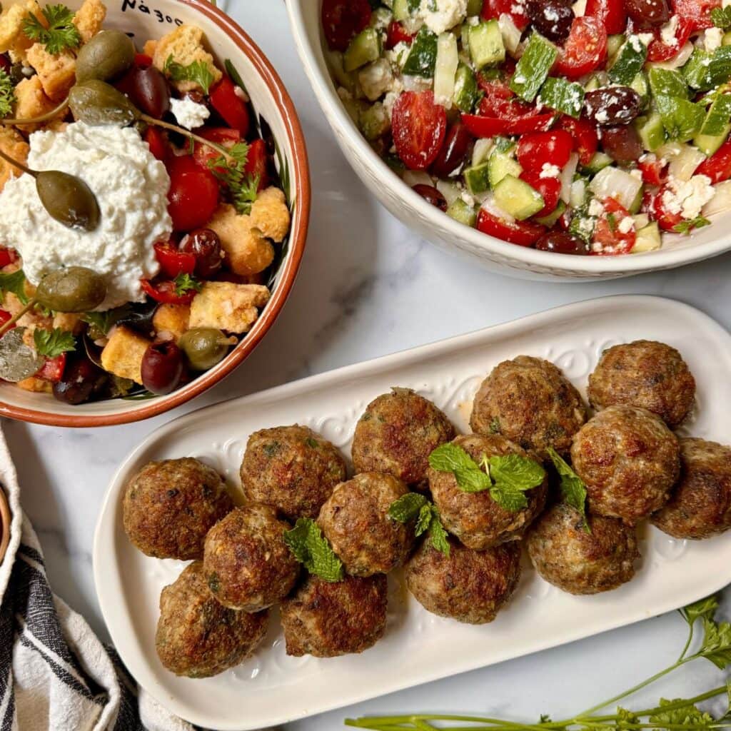 Meatballs and two salads on table with caper berries and cherry tomatoes.