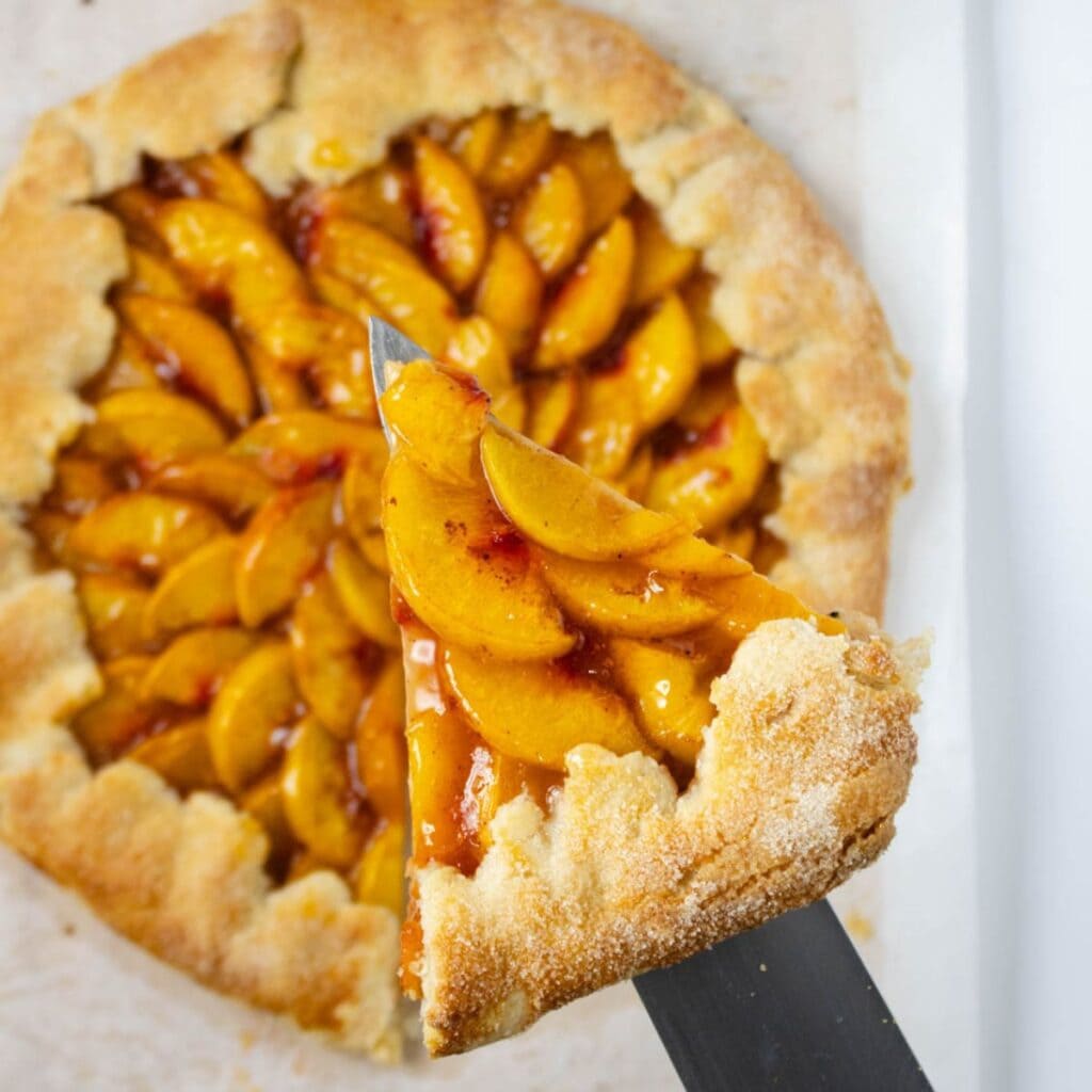 Baked peach galette on parchment paper with one slice pulled out and held above on a knife.