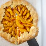 Baked peach galette on parchment paper with one slice pulled out and held above on a knife.