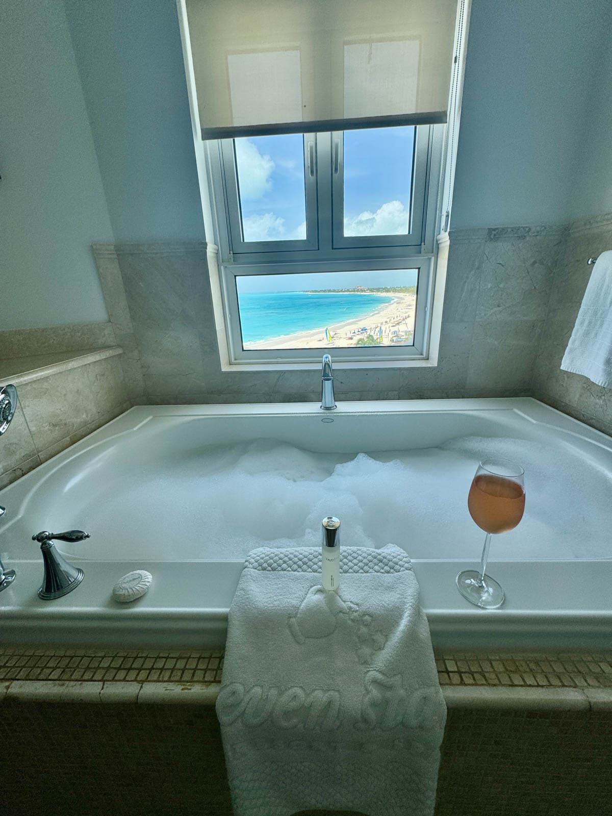 Bath tub with bubbles and towel on side with glass of wine and beach view out the window.
