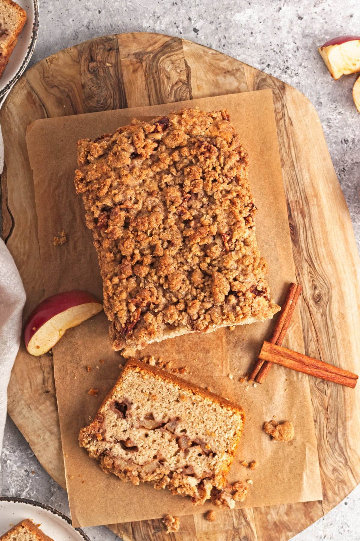 Loaf cake with chopped apples and spices on a wood cutting board with apple slices and cinnamon sticks on a grey counter.