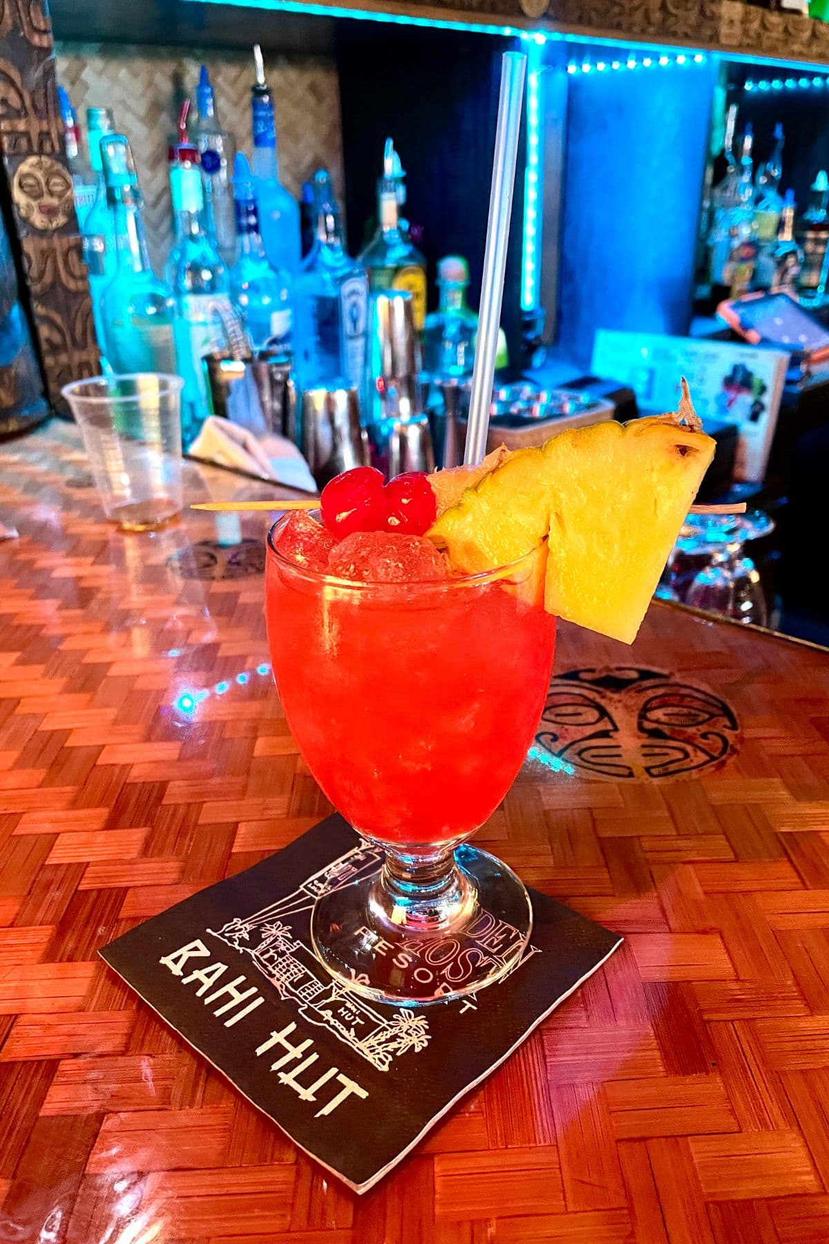 Red cocktail with a slice of pinepapple and cherries on a bar.