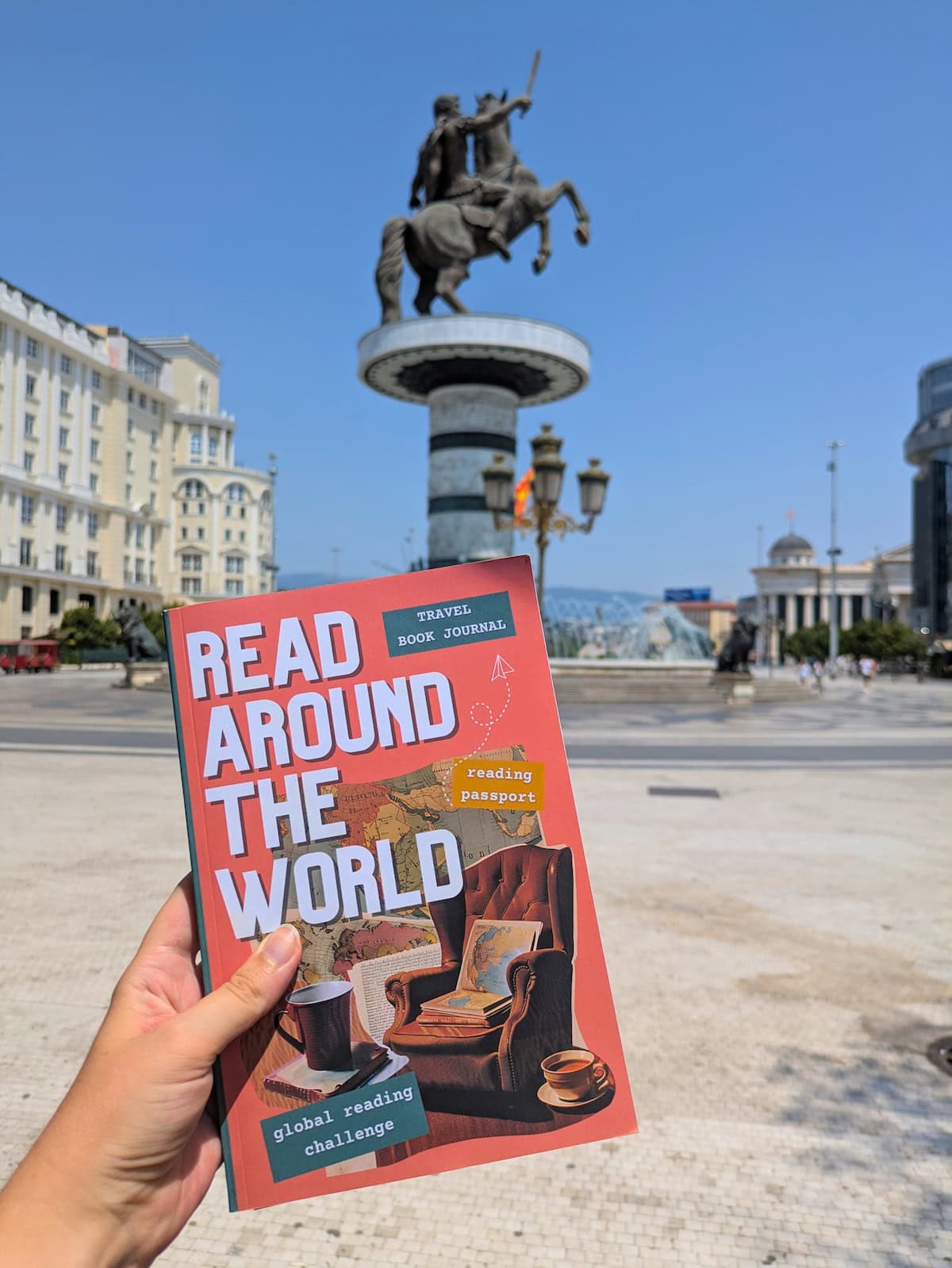 Book in front of a statue against a blue sky with buildings in the background.