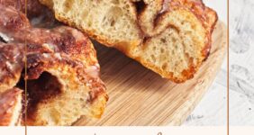 Baked cinnamon and sugar focaccia bread with icing in slices on wood cutting board with blue napkin and cinnamon sticks.