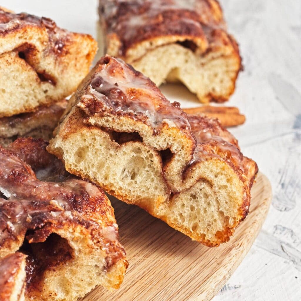 Baked cinnamon and sugar focaccia bread with icing in slices on wood cutting board with blue napkin and cinnamon sticks with icing pitcher on side.