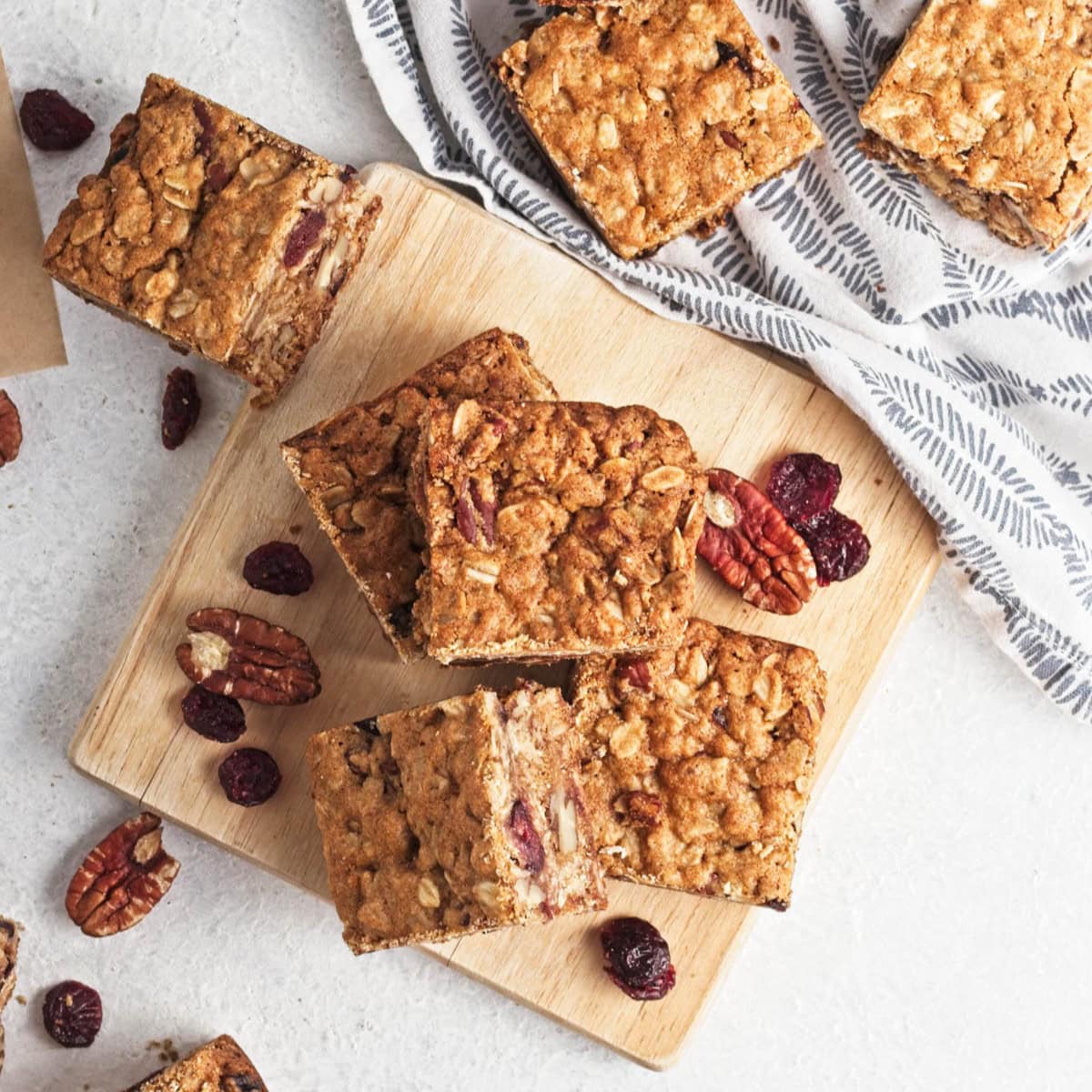 Oatmeal bar squares on cutting board on white counter with some on dish towel and cranberries and pecans scattered.
