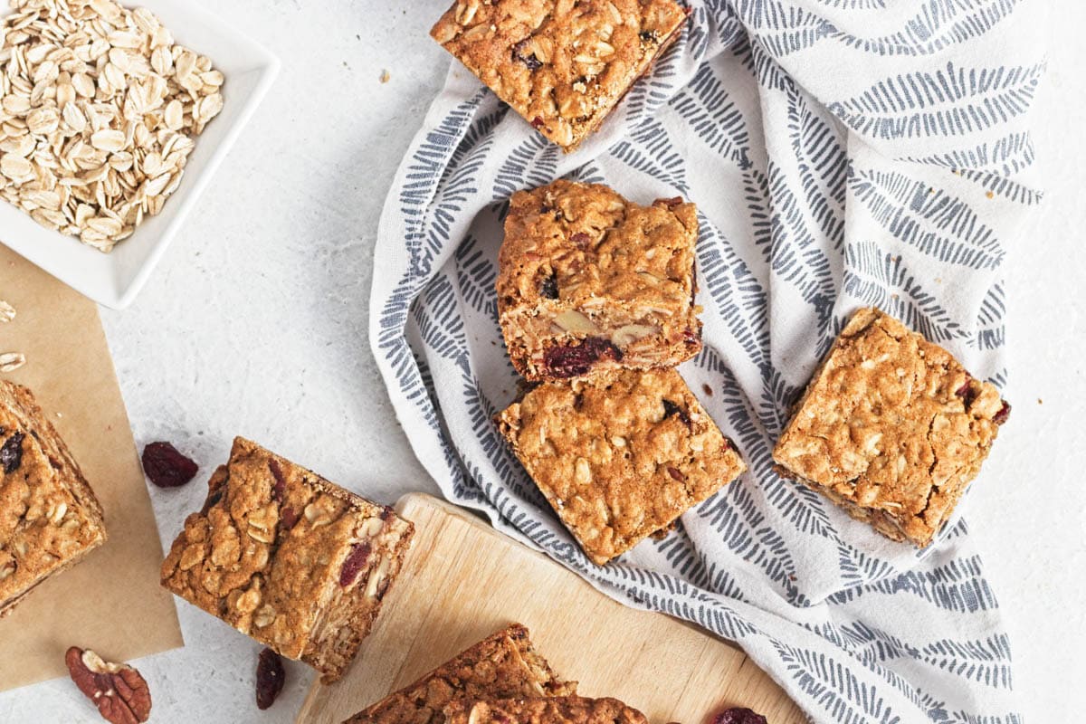Oatmeal bar squares on cutting board on white counter with some on dish towel and cranberries and pecans scattered.