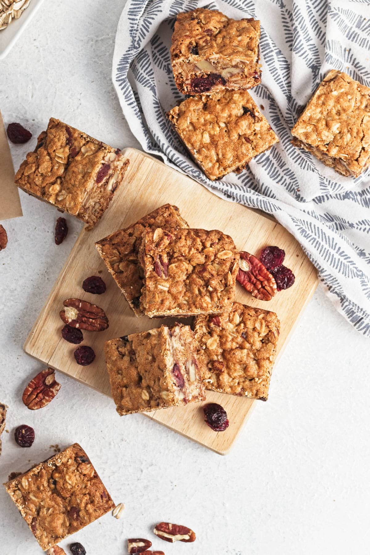 Oatmeal bar squares on white counter with some on cutting board and cranberries and pecans scattered and bars on dish towel in corner.