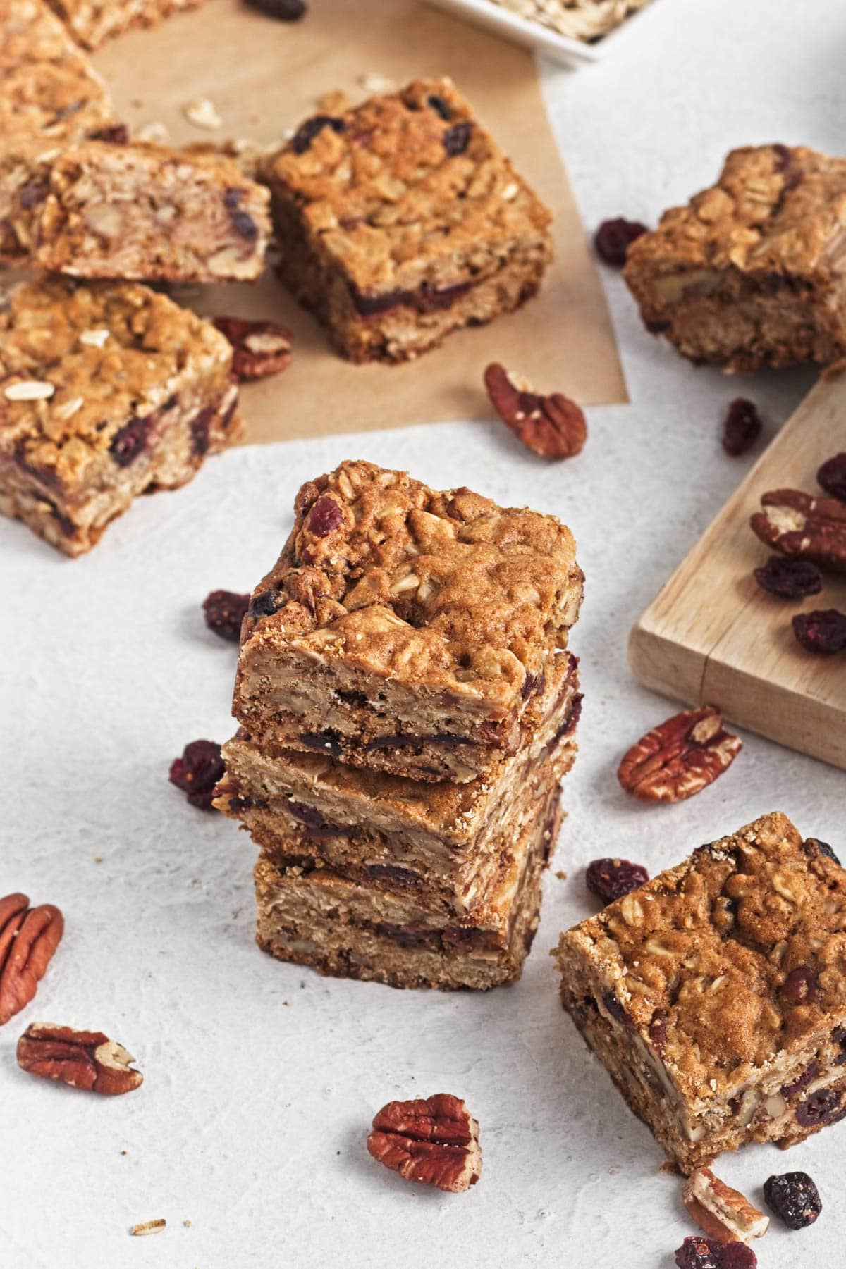 Oatmeal bar squares on cutting board on white counter with some on dish towel and cranberries and pecans scattered.