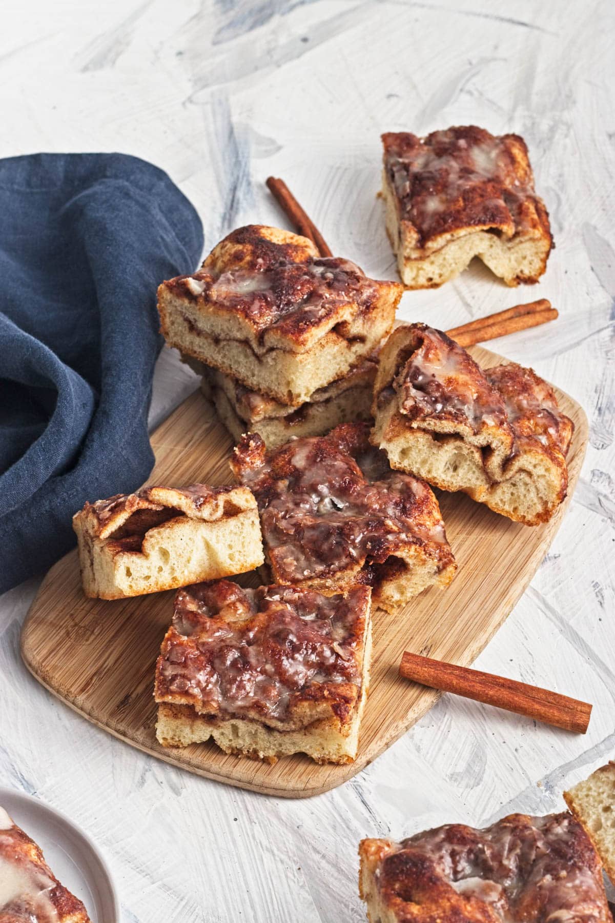 Baked cinnamon and sugar focaccia bread with icing in slices on wood cutting board with blue napkin and cinnamon sticks.