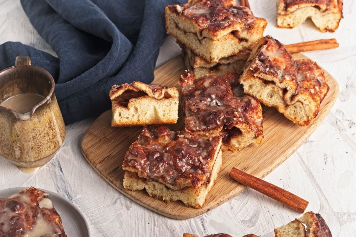 Baked cinnamon and sugar focaccia bread with icing in slices on wood cutting board with blue napkin and cinnamon sticks with icing pitcher on side.