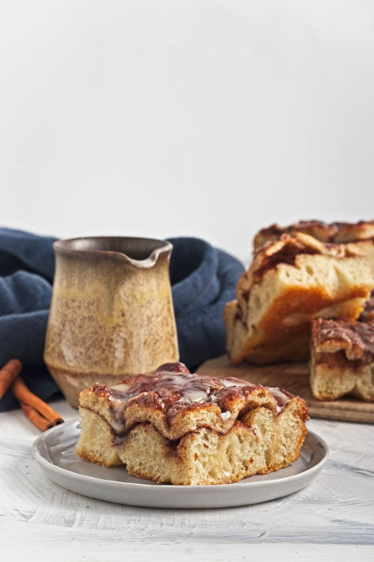 Baked cinnamon and sugar focaccia bread with icing on grey plate with more slices on wood cutting board with blue napkin and cinnamon sticks.