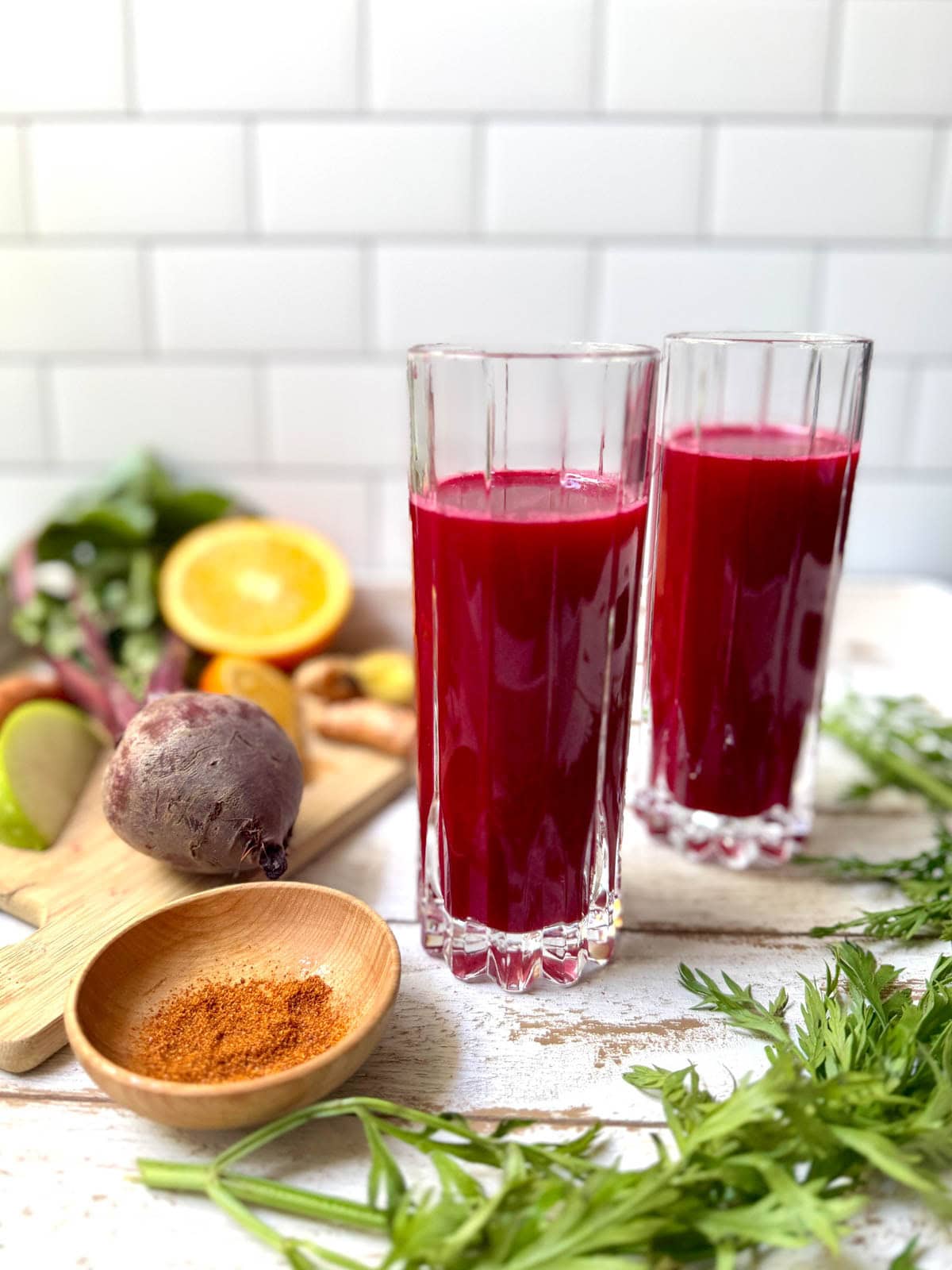 Glasses of fruit and vegetable juice with fruits and vegetables in background and bowl of cayenne pepper.