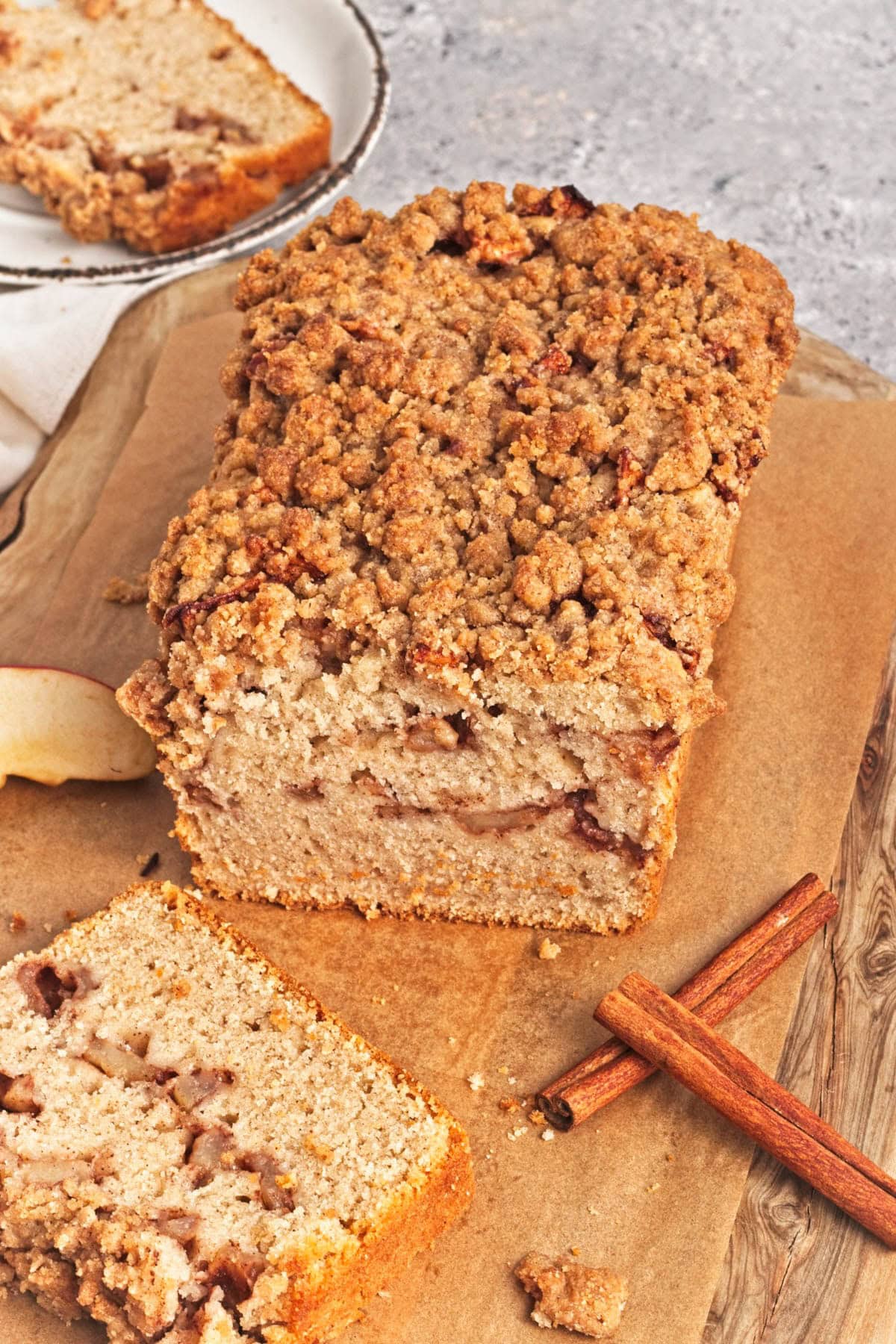 Loaf of apple cake with streusel with slice in front and cinnamon sticks on wood cutting board.