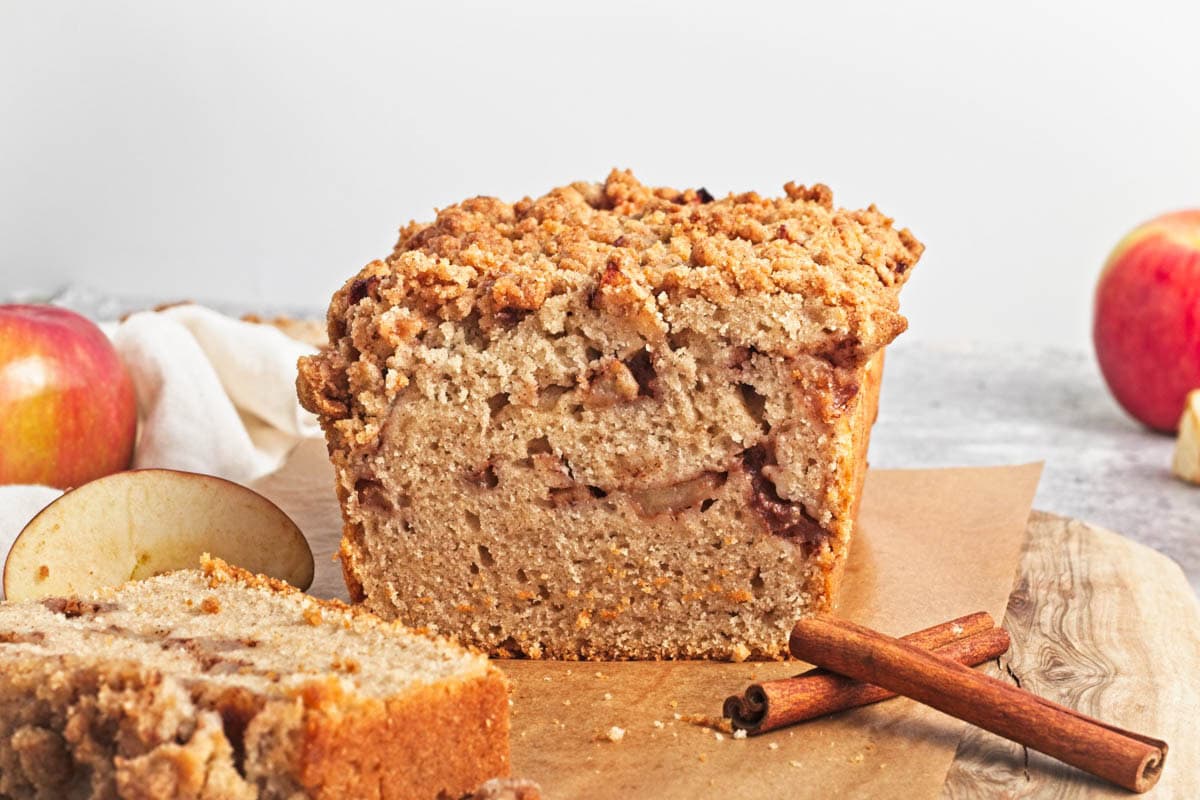 Loaf of apple cake with streusel with slice in front and cinnamon sticks on wood cutting board.