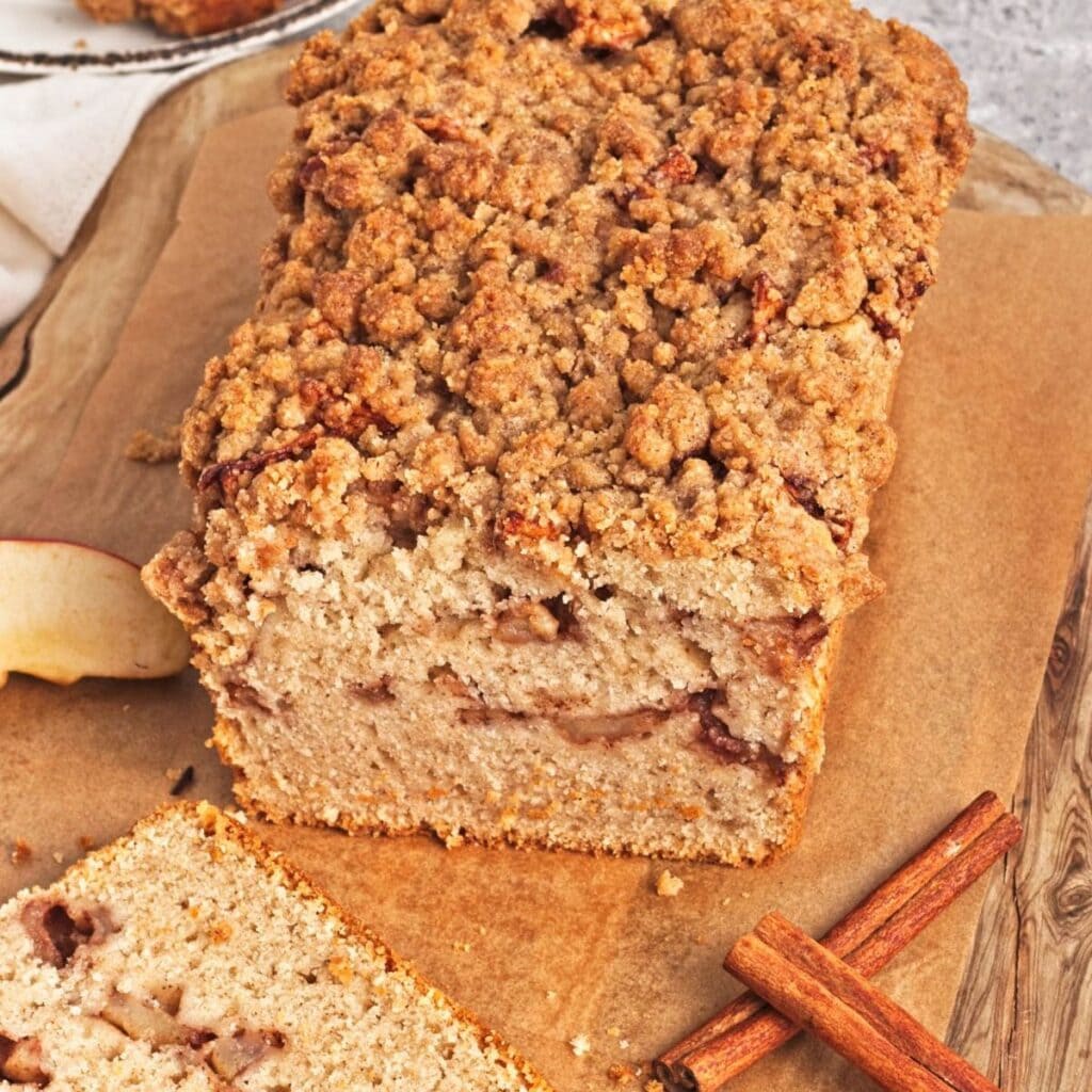 Loaf of apple crumb cake with slice in front and cinnamon sticks on wood cutting board.