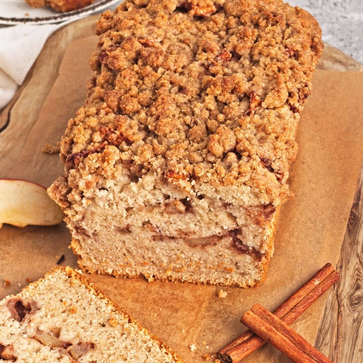 Loaf of apple crumb cake with slice in front and cinnamon sticks on wood cutting board.
