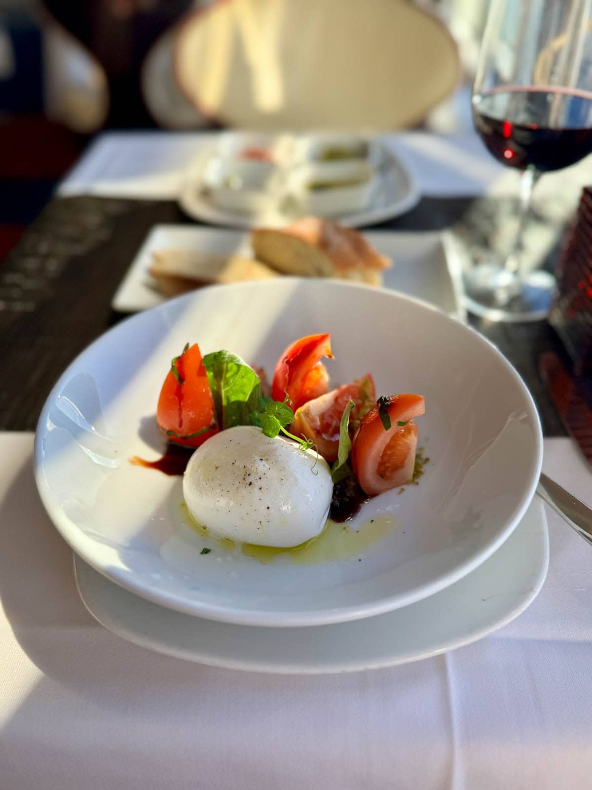 Buratta cheese, tomatoes, and basil with oil and balsamic vinegar in a white bowl with bread and glass of wine in background.
