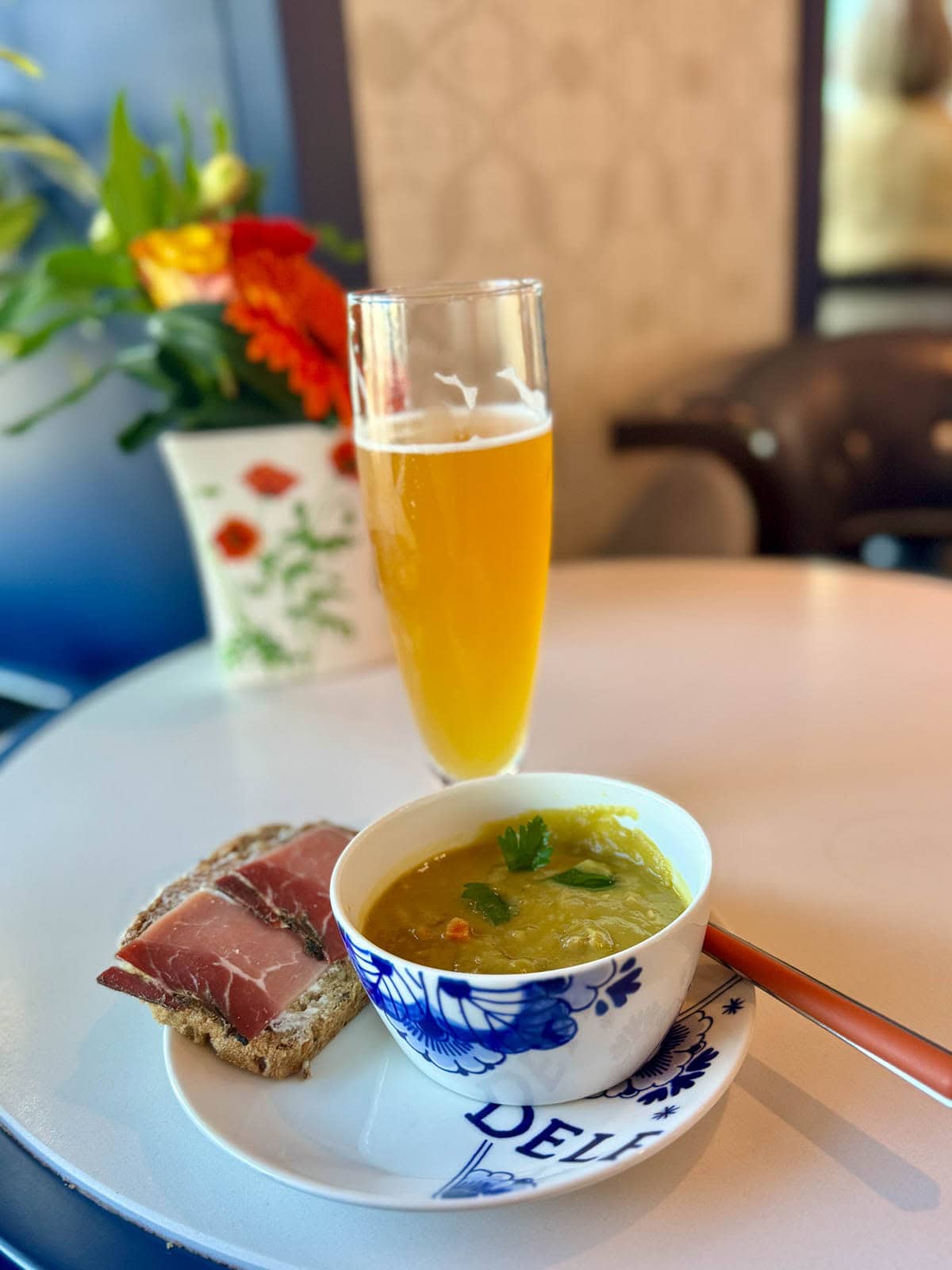 Pea soup with bread, butter, and speck ham on a white and blue plate with a draft beer in background.