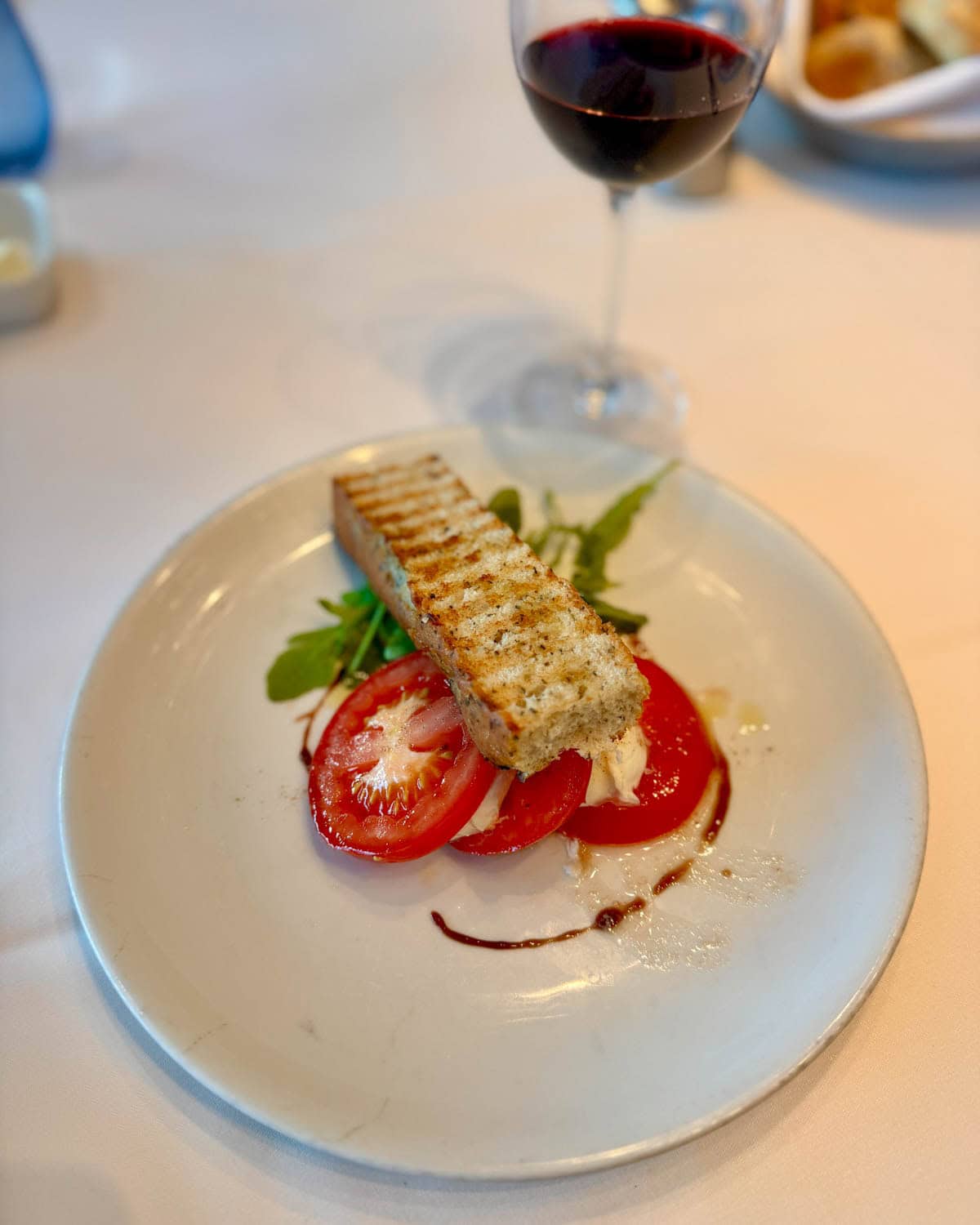 Cheese, tomato, and basil with piece of toasted bread on white plate with glass of red wine in the background.