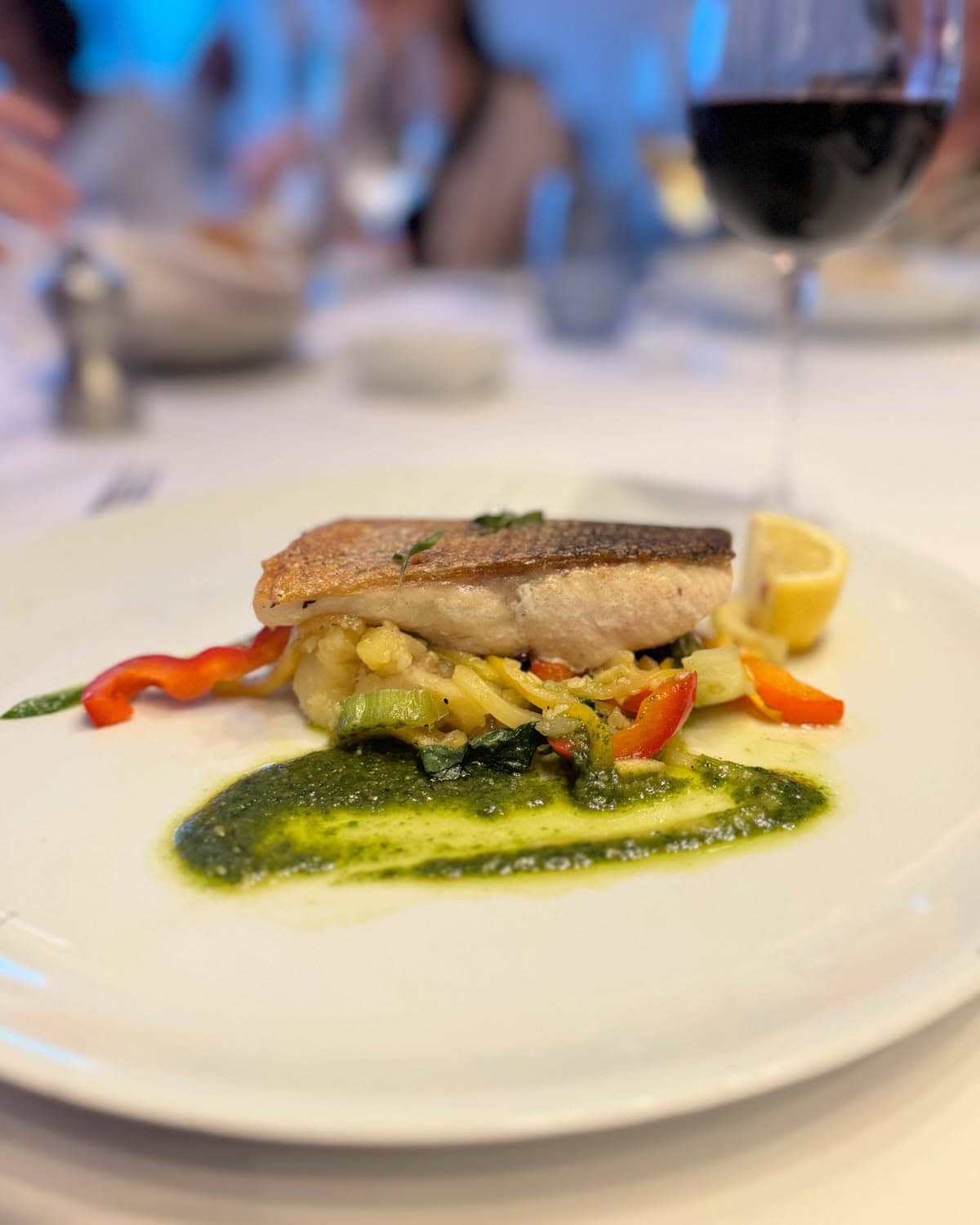 Piece of fish over vegetables with basil sauce, piece of lemon, and glass of wine in background.