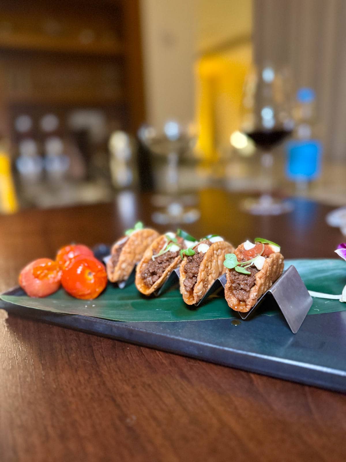Mini tacos with salsa and herbs with side of roasted tomatoes placed on a wood bar.
