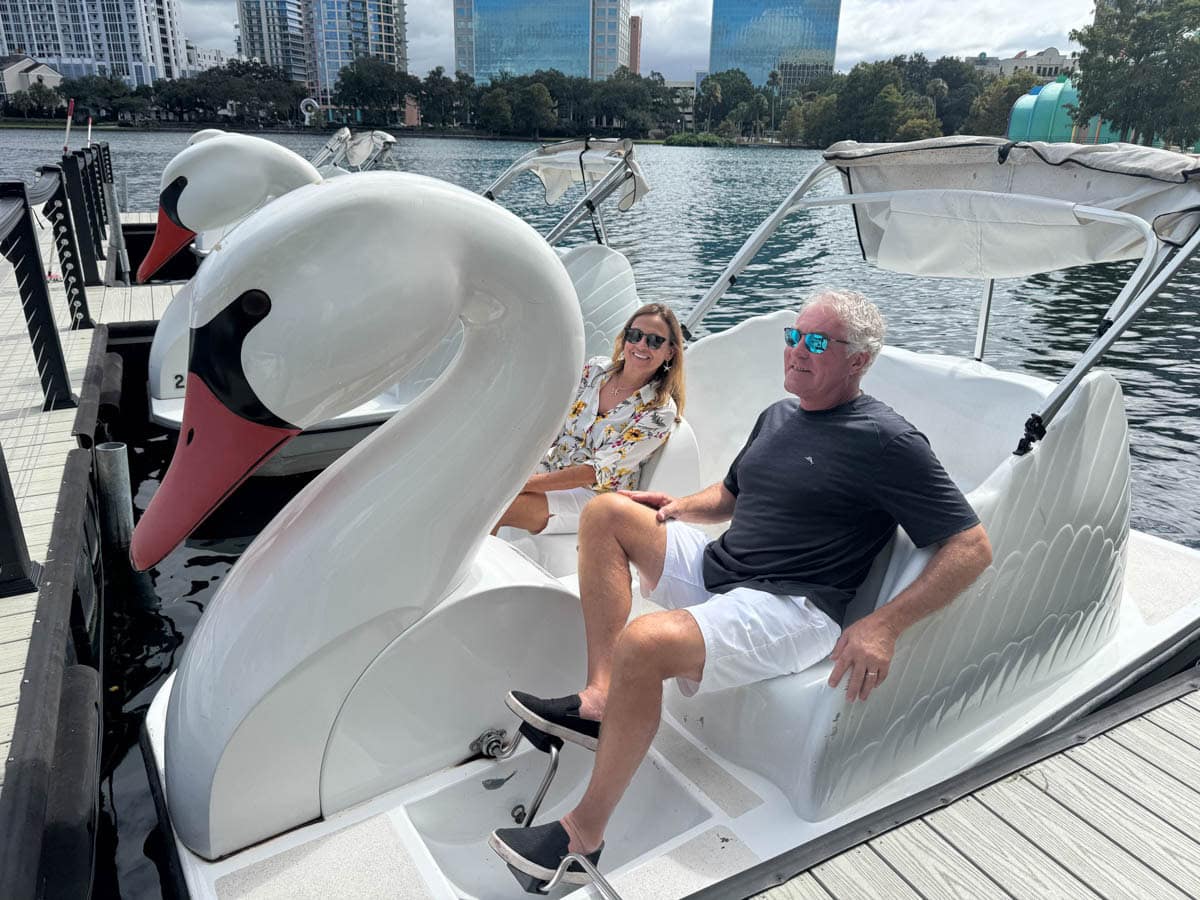 Man and woman in a swan-themed paddleboat with a lake in the background against a blue sky.