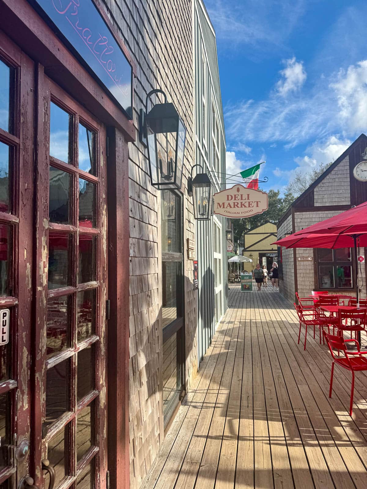 Wood walkway with stores on one side and red tables with unbrellas on the other side.