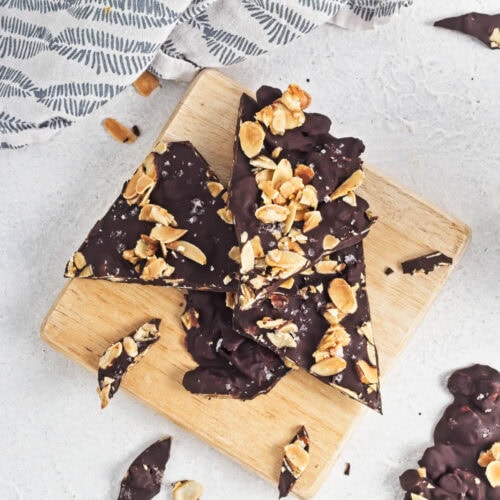 Dark chocolate almond bark on a wood board on a white counter with a dishtowel in the background.