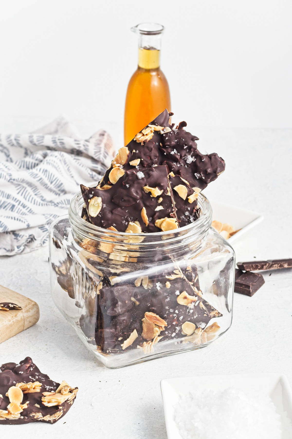 Dark chocolate almond bark in a wide-mouth jar with a bottle of caramel-colored liquid in the background and more bark on a white counter with a dishtowel in the background.