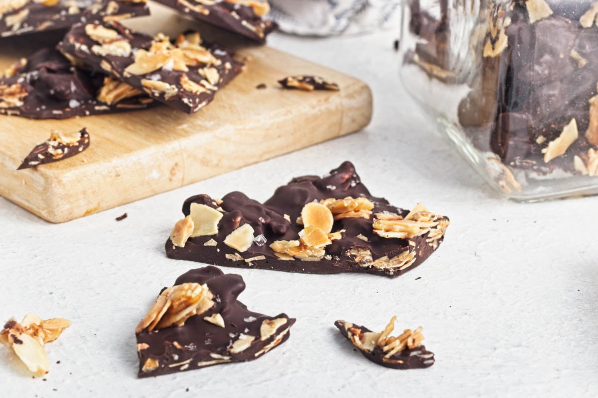 Dark chocolate almond bark on a wood board on a white counter with a dishtowel in the background and more bark in the front.