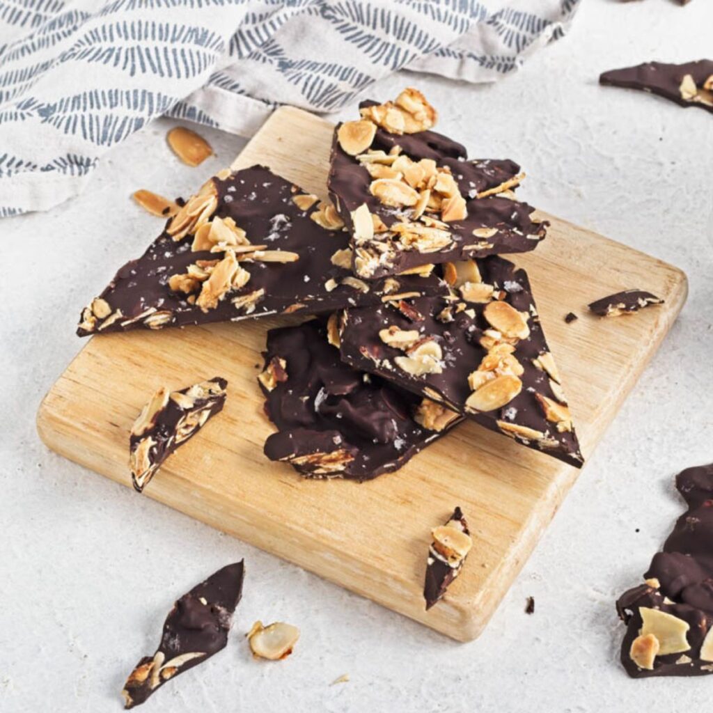 Dark chocolate almond bark on a wood board on a white counter with a dishtowel in the background.