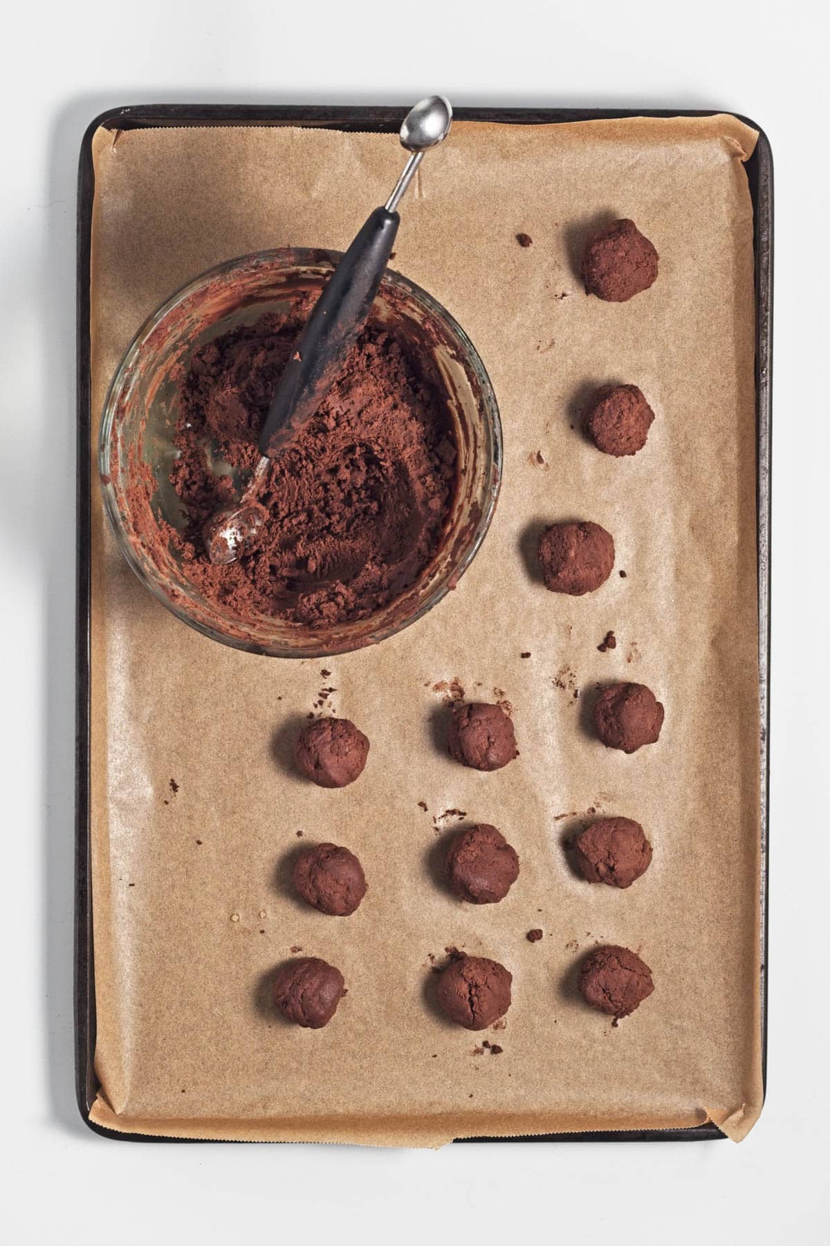Chocolate balls and glass bowl of chocolate with a small scoop on parchment paper on baking sheet.