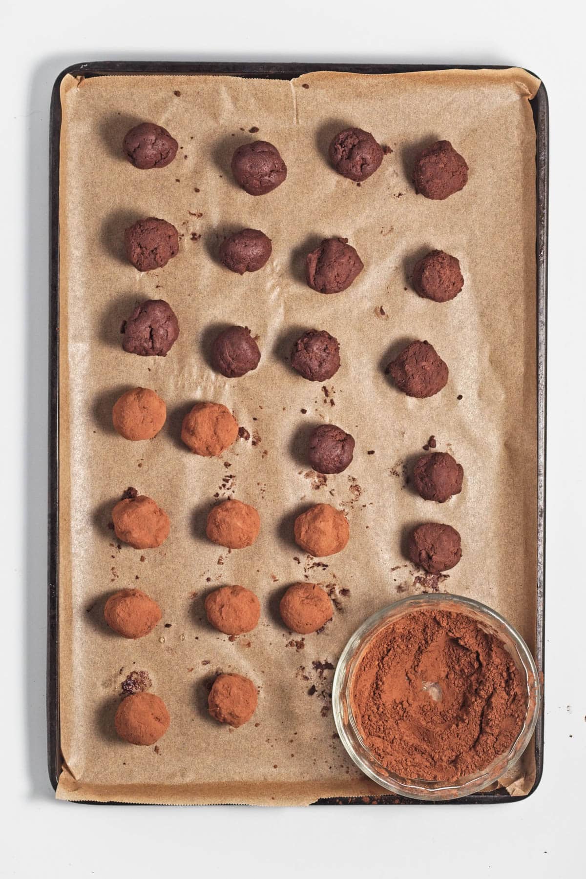 Chocolate balls, some rolled in cocoa powder, and a glass bowl of chocolate with a small scoop on parchment paper on baking sheet.