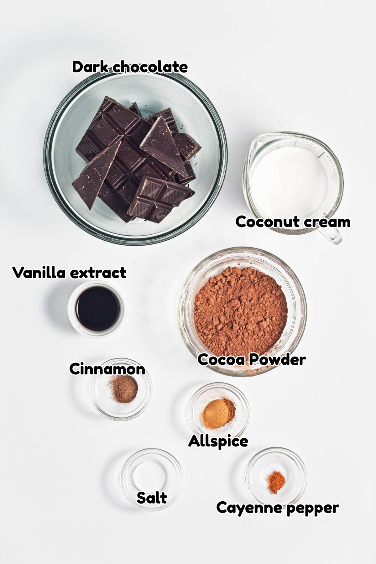 Chocolate, coconut cream ,cocoa powder, vanilla extract, cinnamon, allspice, cayenne pepper, and salt in glass bowls on a white counter