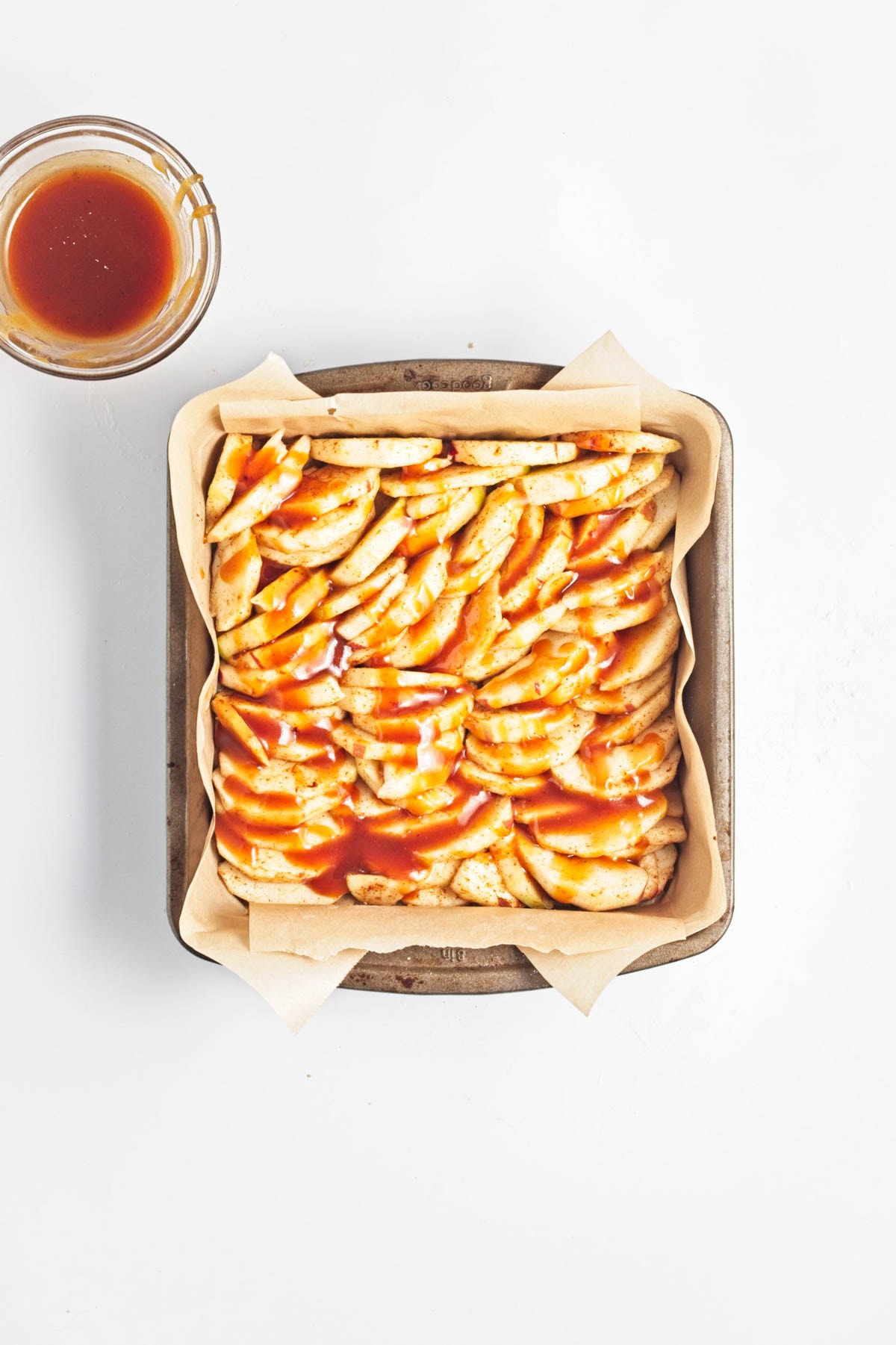 Sliced apples in a parchment paper-lined baking dish with caramel sauce drizzled on top.