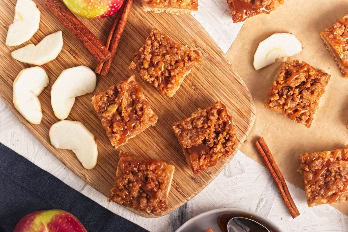 Fruit bars with crumble and caramel on top and apple slices and cinnamon sticks around on wood board and parchment.