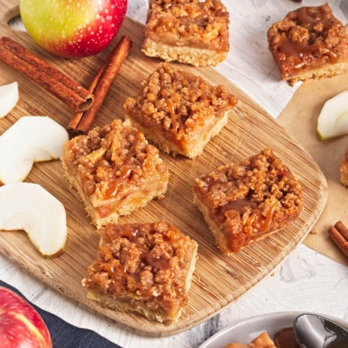 Fruit bars with crumble and caramel on top and apple slices and cinnamon sticks around on wood board and parchment paper.