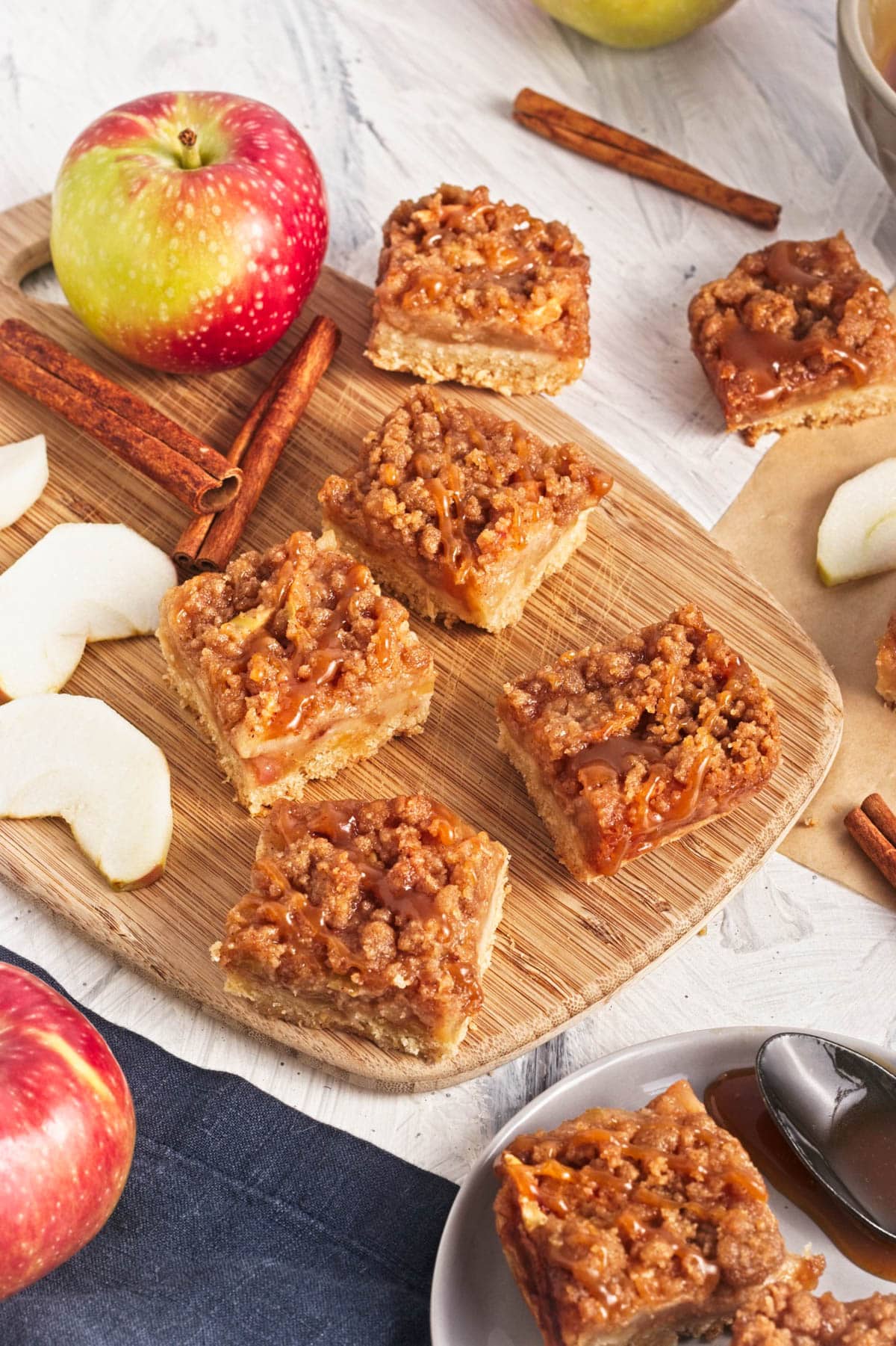 Fruit bars with crumble and caramel on top and apple slices and cinnamon sticks around on wood board and parchment paper.