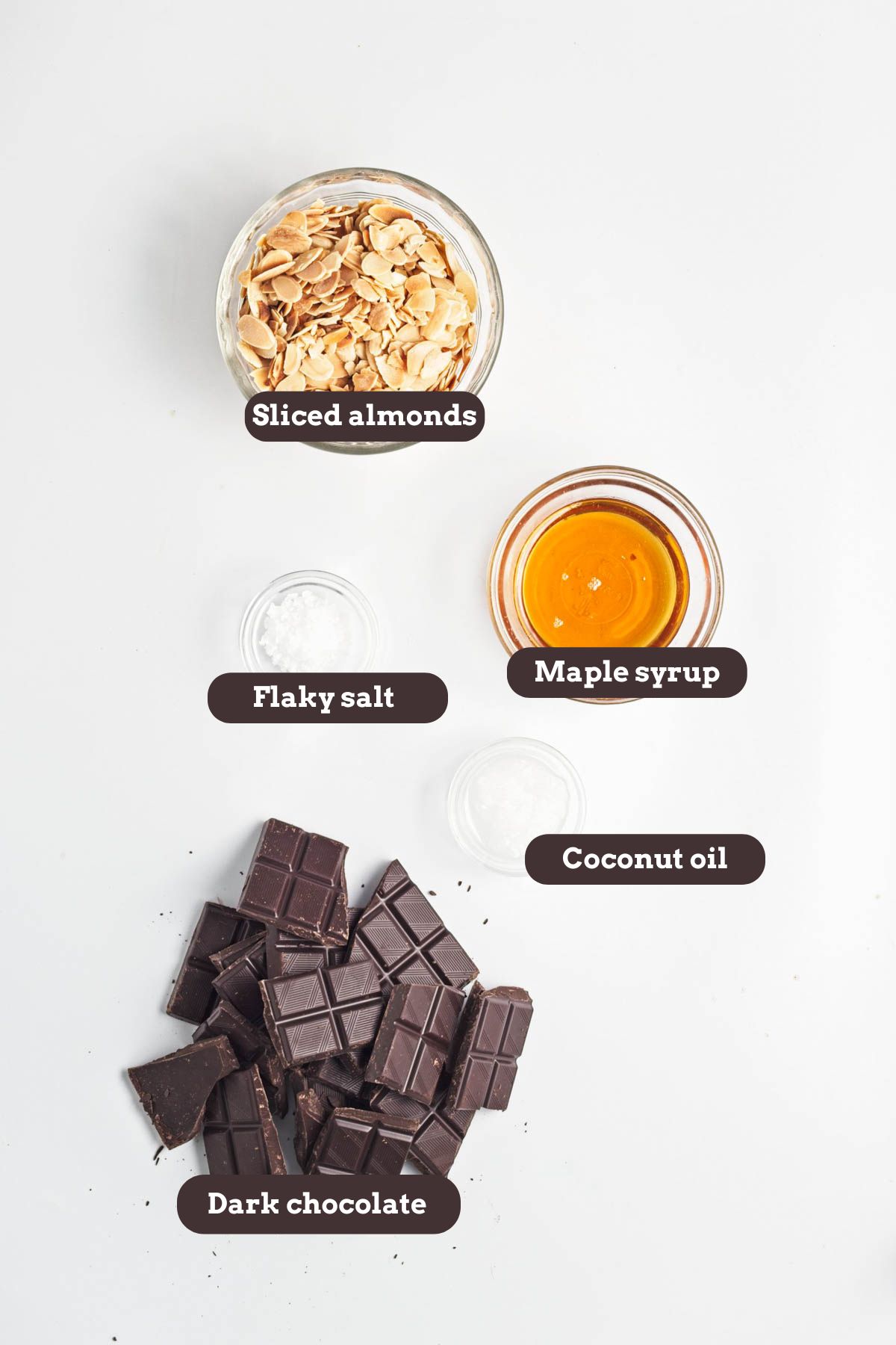 Sliced almonds, maple syrup, coconut oil, salt, and ark chocolate on a white coiunter.