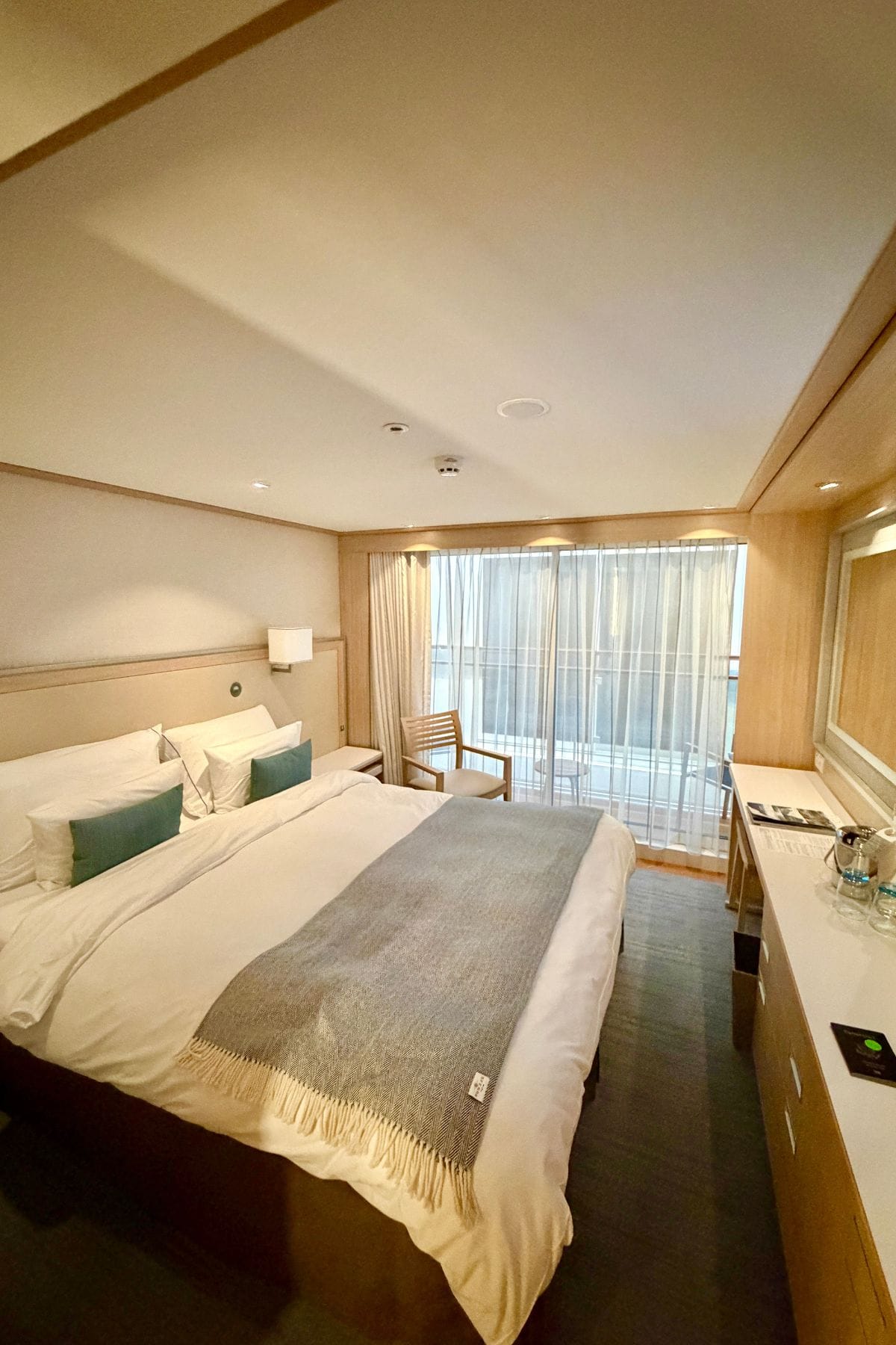 Cruise cabin with bed, counter, and chair in front of a draped window.