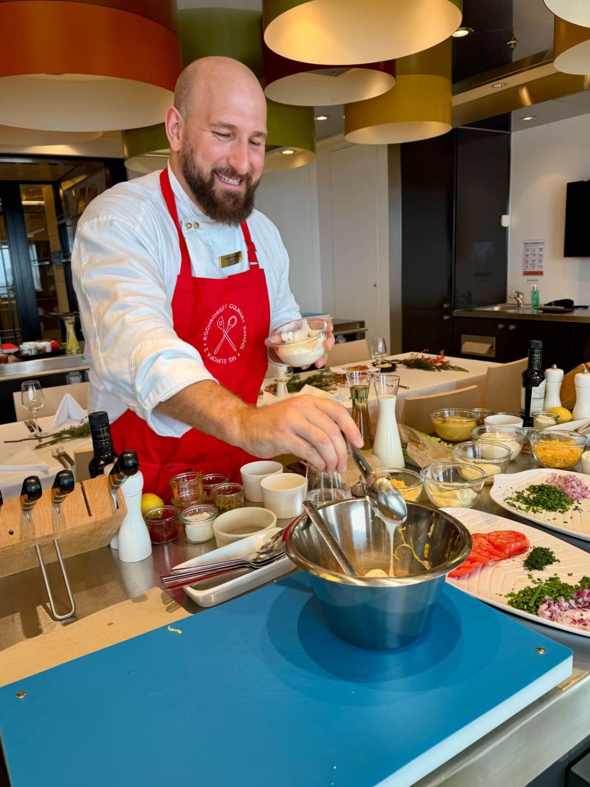 Man in red apron spooning a white substance into a stainless bowl next to plates with tomatoes, parsley and onions.