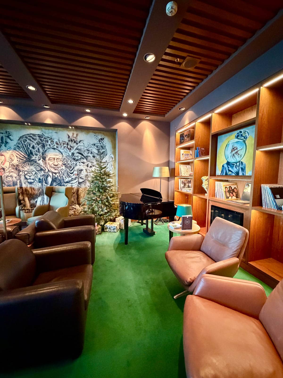 Lounge with green carpet, leather chairs, a grand piano, bookshelves, and art work on the back wall.