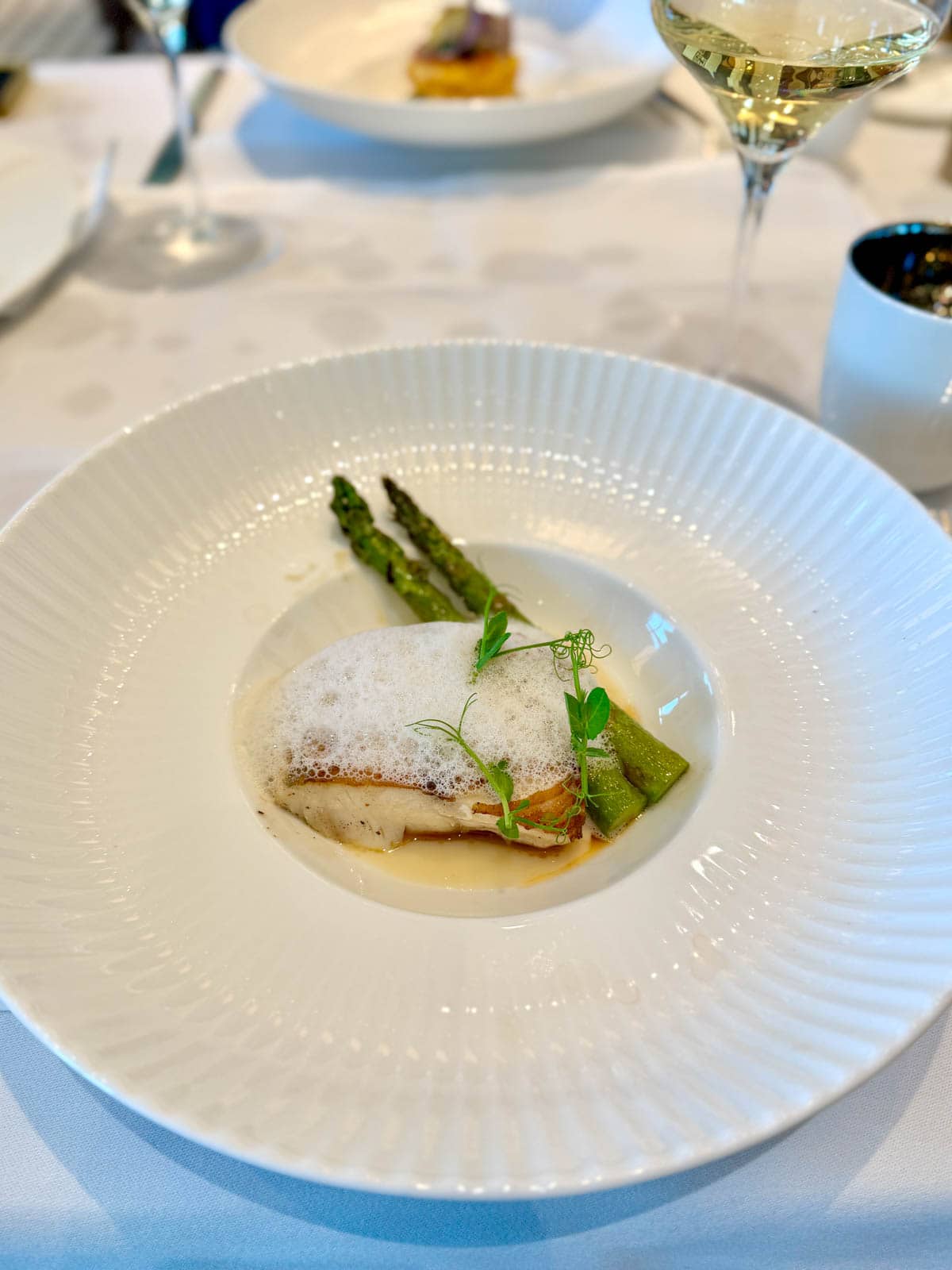 Fish topped with foam with asparagus in a white bowl.