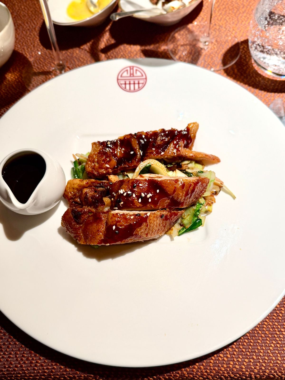 Sliced grilled duck with dark sauce on top with small pitcher to the side on a white plate.