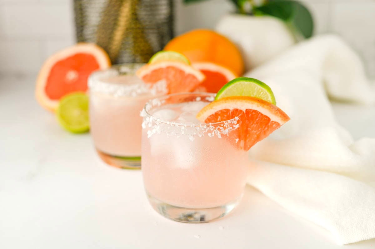 Two glasses containing pink liquid with salt on rims, fruit slices on rim, and white napkin and plant background.