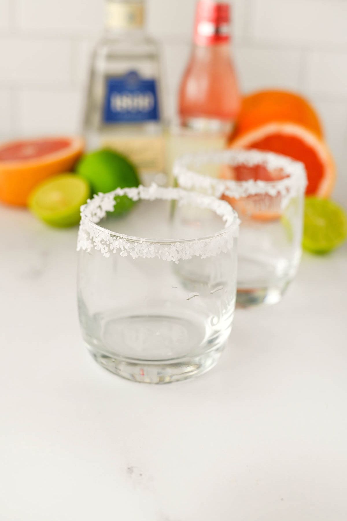 Two glasses with salt on rims and bottles and fruit in background.