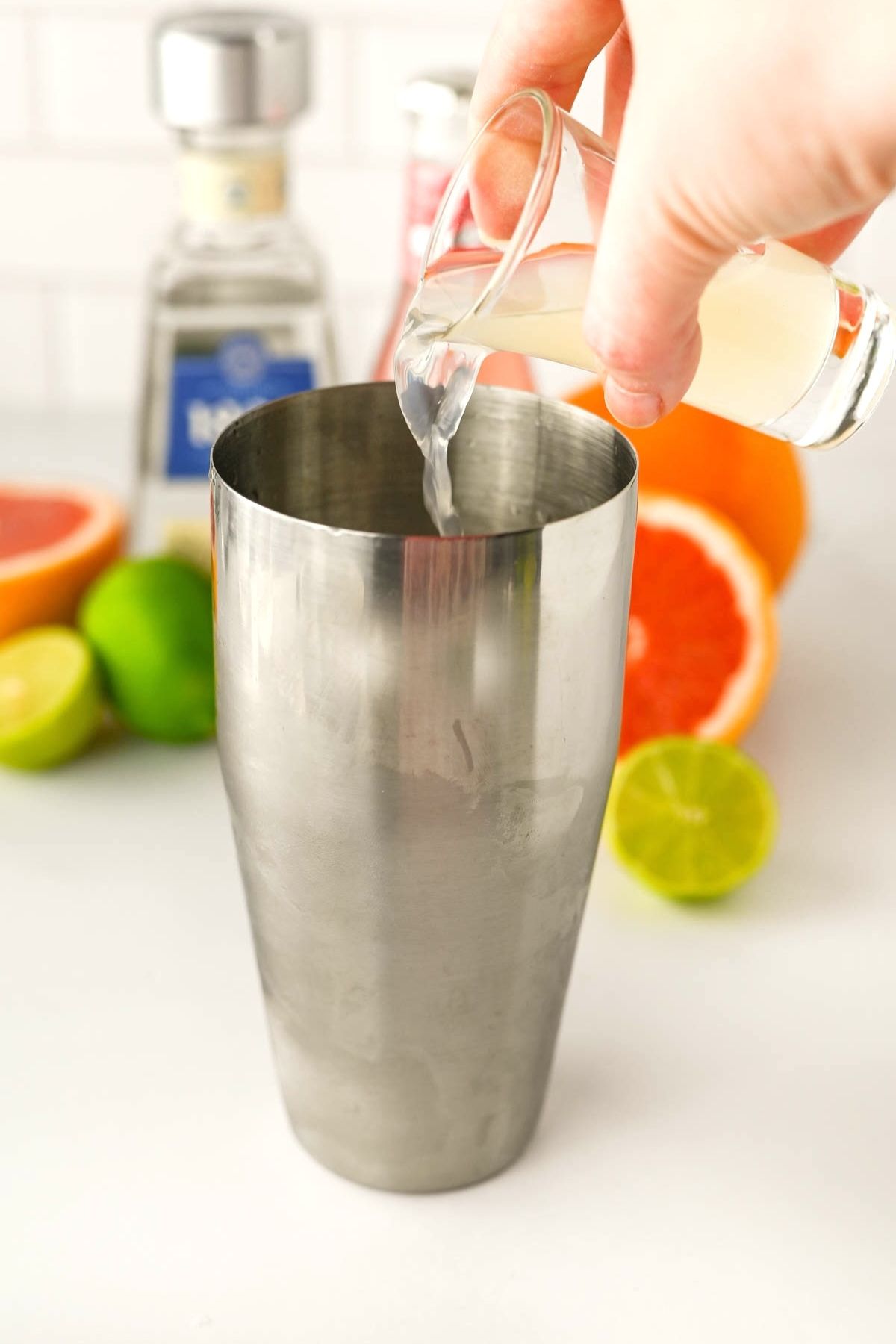 Stainless shaker with hand pouring yellow liquid into shaker, with bottle and fruit in background.