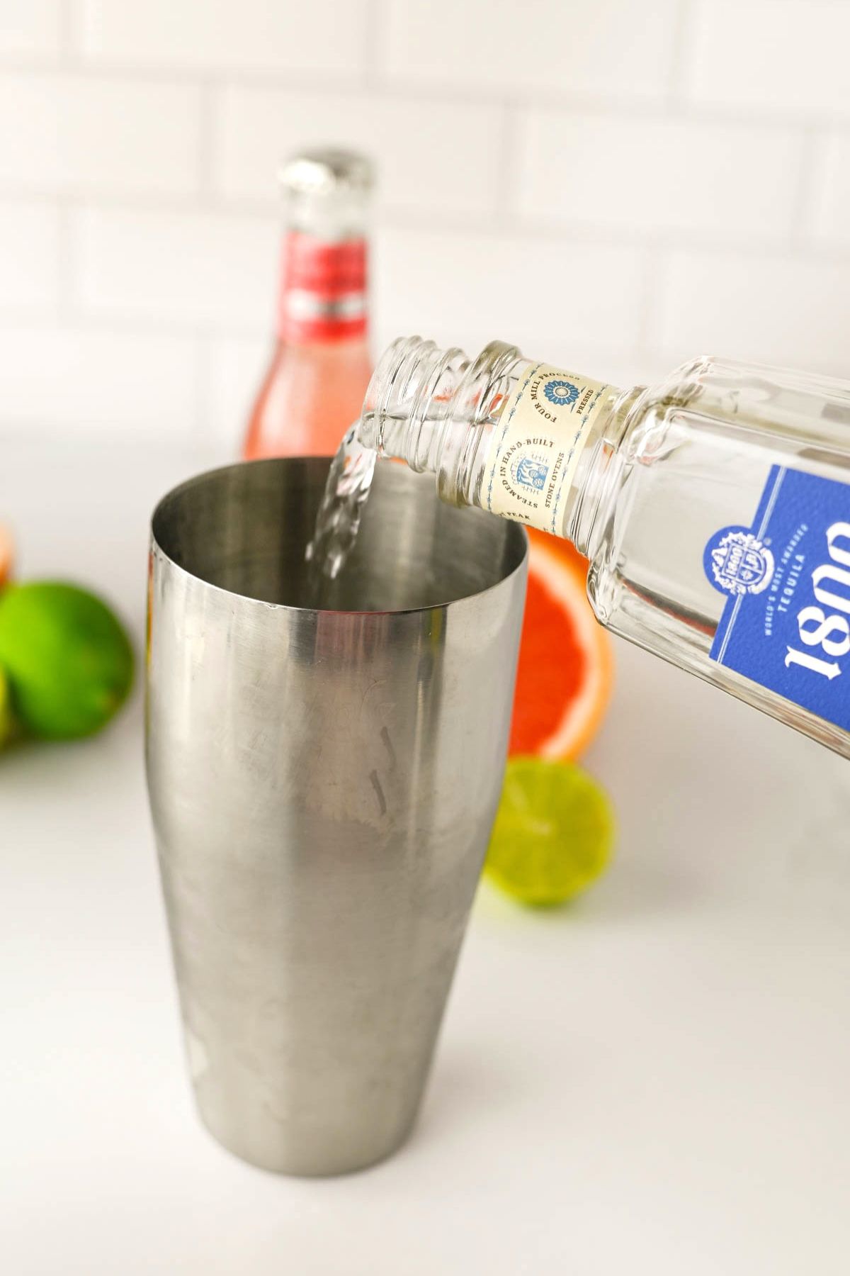 Stainless shaker with hand pouring clear liquid into shaker, with bottle and fruit in background.