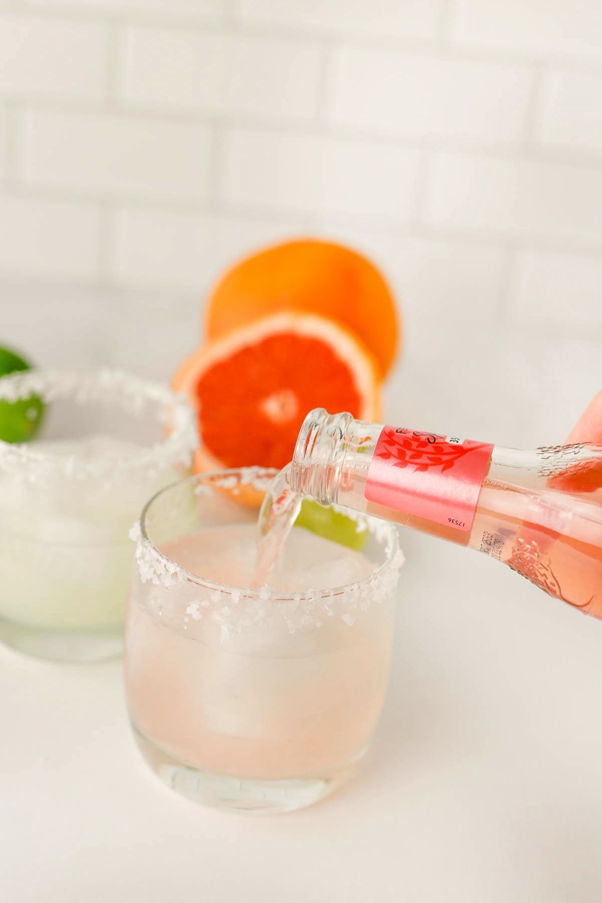 Two glasses with salt on rims and yellow liquid with hand pouring pink liquid into front glass.